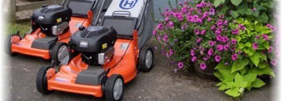 Two orange Husqvarna push mowers parked on gravel beside a planter of purple flowers, the banner photograph from the company's own 2022 website